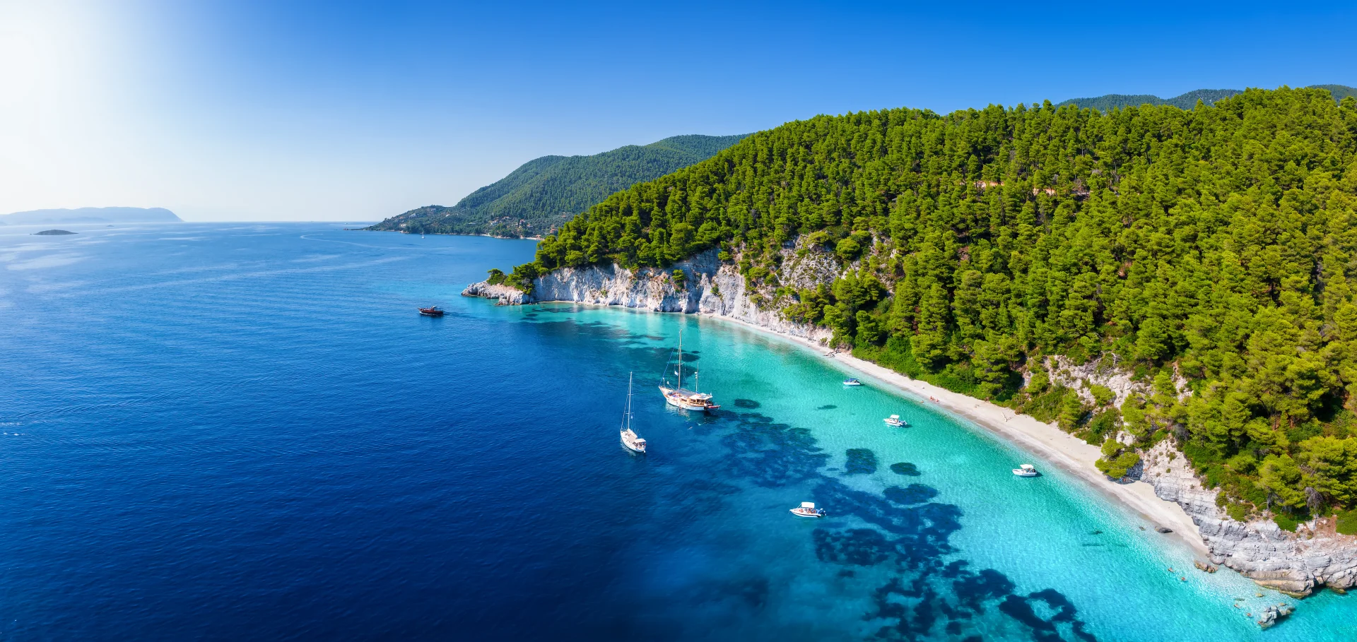 Peaceful pine-covered coastline with secluded beaches and yachts anchored in turquoise waters.