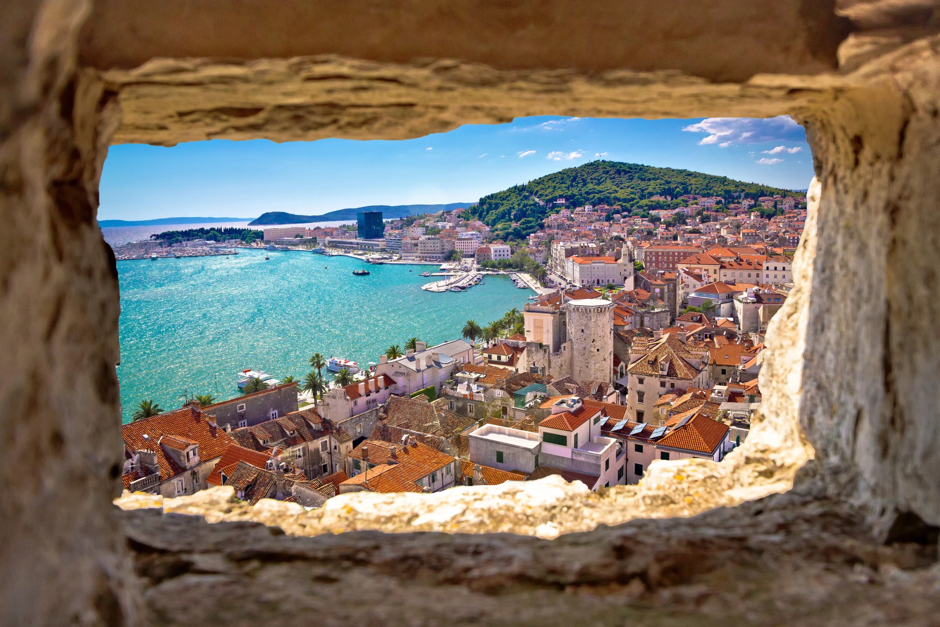 A picturesque view of Split's historic terracotta rooftops and vibrant harbor, perfectly framed through an ancient stone window.